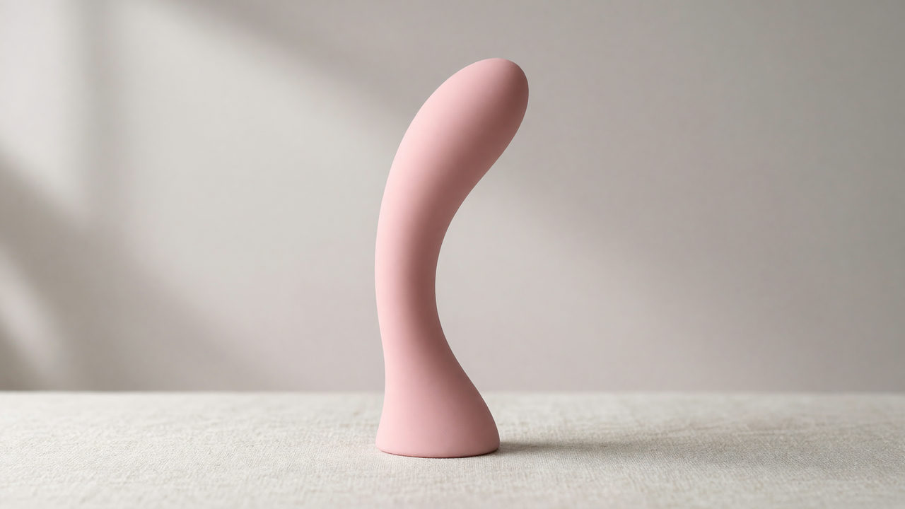 Een roze siliconen vibrator met een gebogen schacht en een brede afgeronde kop, rechtop op een lichte ondergrond.